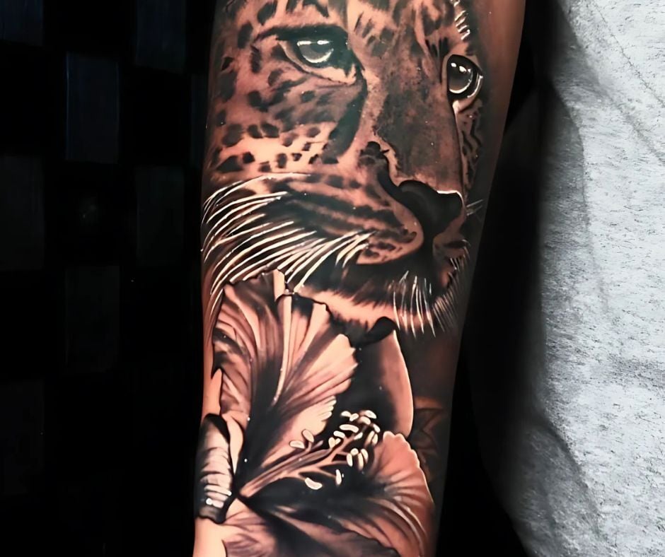 Realism forearm tattoo of a leopard with lifelike eyes and a hibiscus flower, high-contrast black-and-grey shading.